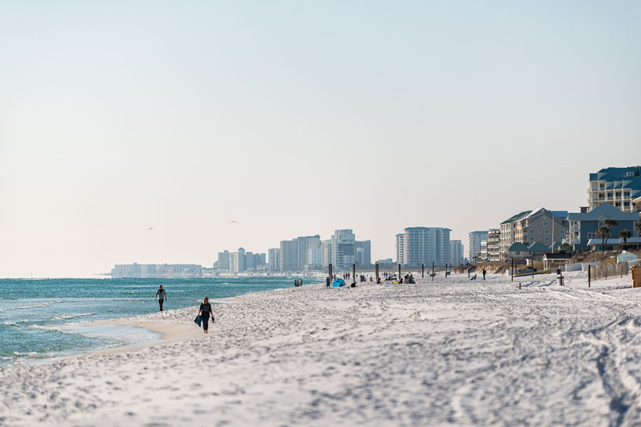 Visitors walk along the sugar-white sand shoreline of Miramar Beach, with Emerald Coast high-rise resorts and condos stretching along the Gulf in the distance.