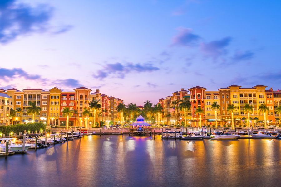 Evening view of Bayfront Naples in Naples, Florida, featuring waterfront residences, restaurants, and marinas along the Gordon River, with illuminated buildings and boats reflecting the city’s upscale coastal atmosphere.