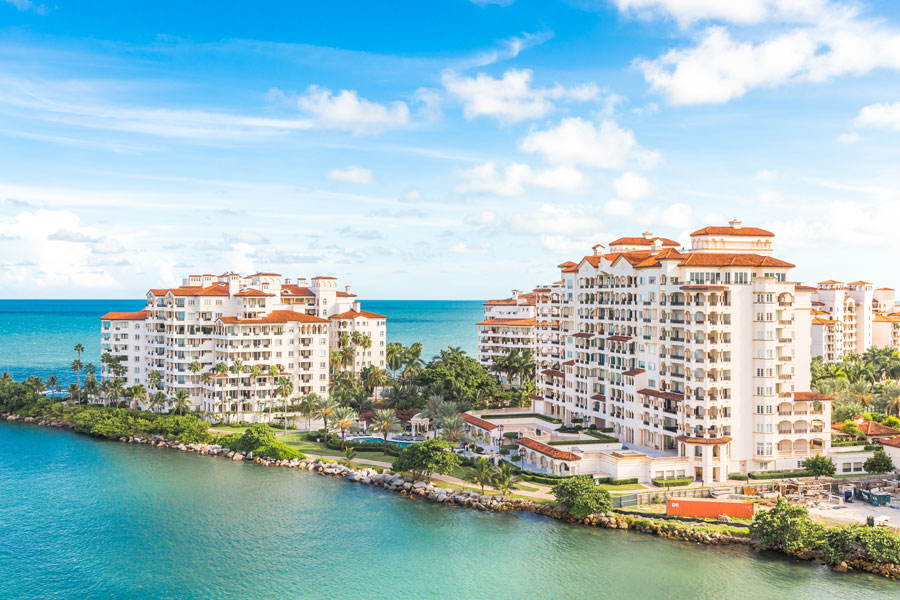 Waterfront residential buildings on Fisher Island, Florida, featuring Mediterranean-style architecture, landscaped grounds, and views of Biscayne Bay and the Atlantic Ocean beyond.