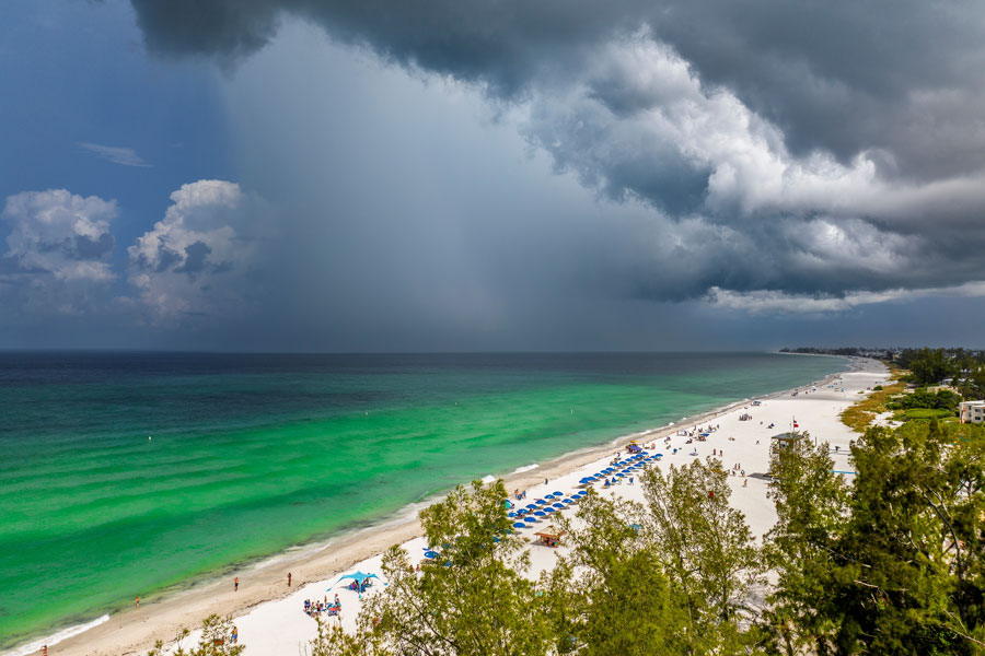 Dark summer storm clouds gather over the turquoise waters and white-sand shoreline of Anna Maria Island along Florida’s Gulf Coast. The scene captures the region’s dramatic subtropical weather, where bright beach conditions can quickly give way to afternoon thunderstorms during the warmer months.