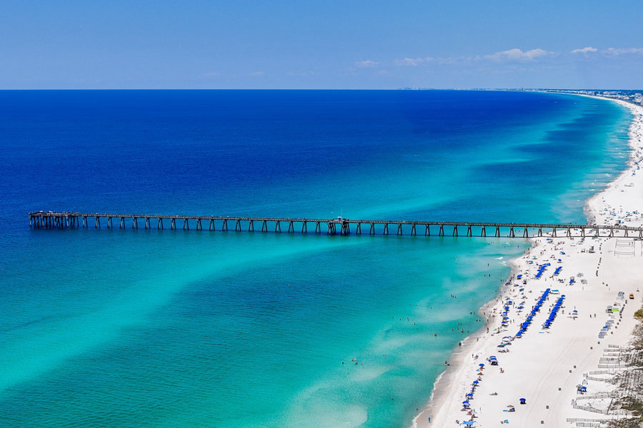 Aerial view of Panama City Beach along Florida’s Panhandle, where a long fishing pier extends over the clear blue-green Gulf waters and bright white sand shoreline. The area is one of Northwest Florida’s best-known beach destinations, with miles of public coastline, resorts, recreation, and easy access to nearby parks and attractions.
