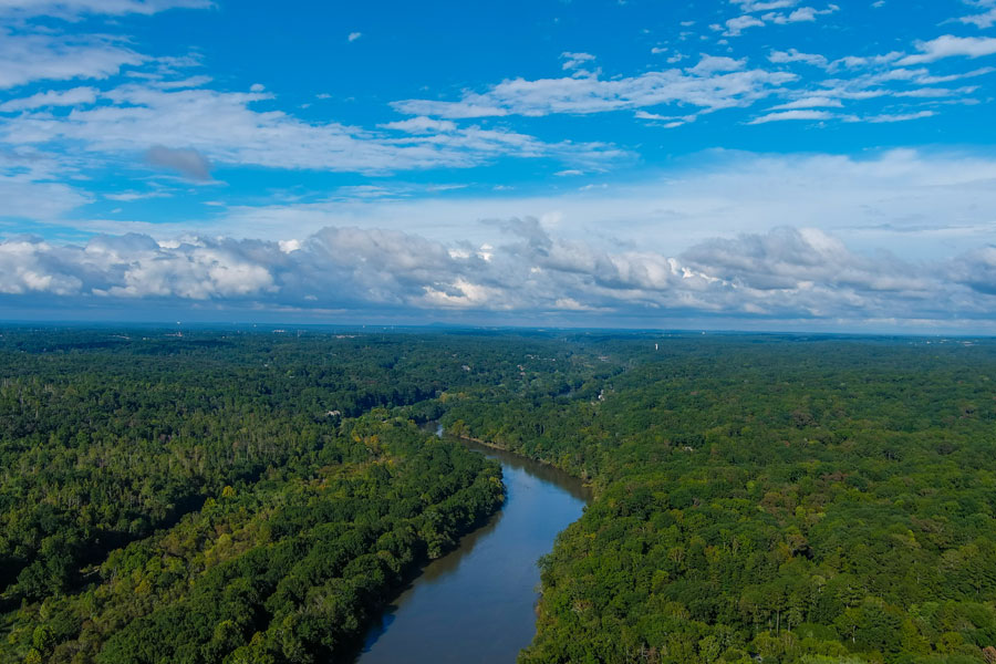An aerial view of the Chattahoochee River winding through a broad forested landscape, with dense tree cover stretching toward the horizon beneath a dramatic blue sky. The river and surrounding woodlands illustrate the expansive natural scenery found along the Chattahoochee watershed.