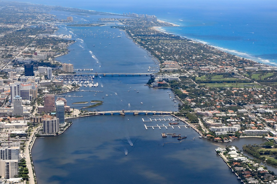 An aerial view of Lake Worth Lagoon looking north between downtown West Palm Beach and the island of Palm Beach, with bridges spanning the lagoon and Singer Island visible in the distance. The expansive view highlights the lagoon’s role as a major waterway separating the mainland from Palm Beach County’s Atlantic barrier islands.