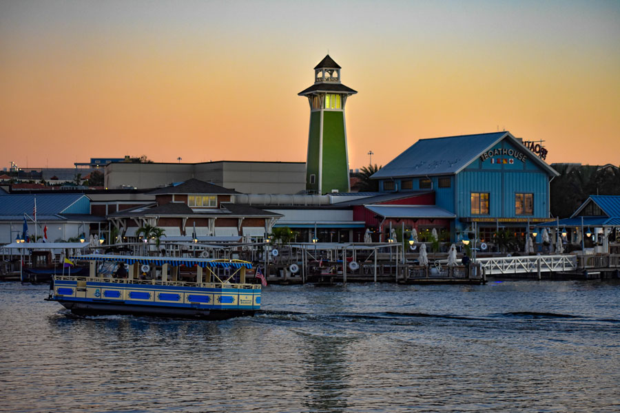 A colorful water taxi cruises across the waterfront at Disney Springs in Lake Buena Vista, Florida, as the sun sets behind the shopping and entertainment district's distinctive lighthouse and waterfront restaurants. Part of Walt Disney World Resort, Disney Springs is a popular Central Florida destination offering waterfront dining, specialty shopping, live entertainment and complimentary boat transportation connecting select Disney resort hotels. Photo credit: VIAVAL TOURS, licensed.