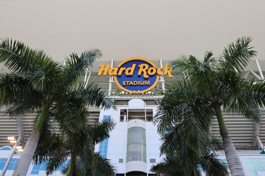 The Hard Rock Stadium sign is prominently displayed above the entrance to the landmark sports and entertainment venue in Miami Gardens, Florida, photographed on March 27, 2019, during the Miami Open tennis tournament. Framed by tropical palm trees, the stadium's distinctive architecture and expansive canopy reflect its role as one of South Florida's premier sporting destinations. Home to the NFL's Miami Dolphins and the University of Miami Hurricanes football team, Hard Rock Stadium also hosts international tennis competitions, major soccer matches, concerts, and other large-scale events. Photo credit: Leonard Zhukovsky; licensed.