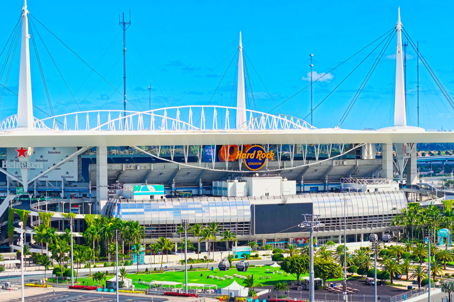 An elevated view of Hard Rock Stadium in Miami Gardens, Florida, showcases its distinctive white canopy roof, towering support structures, and landscaped surroundings. Home to the NFL's Miami Dolphins and the University of Miami Hurricanes football team, the stadium also hosts major international sporting events, concerts, and entertainment attractions. Originally opened in 1987, the venue has undergone extensive renovations that have transformed it into one of South Florida's premier sports and entertainment destinations.