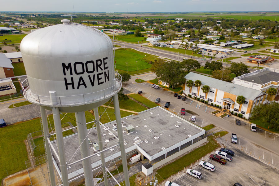 An aerial drone view shows Moore Haven’s municipal water tower rising above the town center and surrounding buildings. The elevated tank is part of the community’s public water system and serves as a prominent landmark in Glades County. Beyond the downtown streets, the flat agricultural landscape extends toward Lake Okeechobee. Photo credit: Felix Mizioznikov, licensed.
