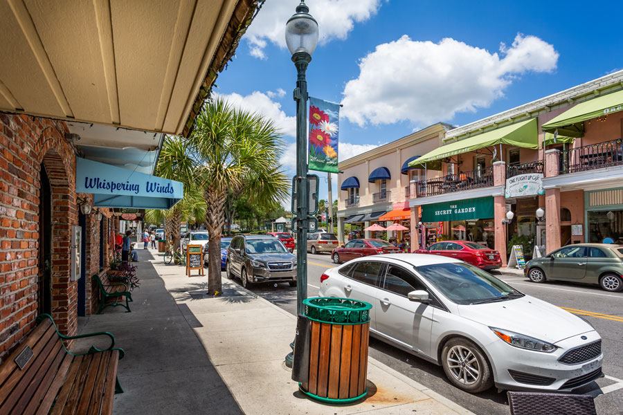 A street-level view of historic downtown Mount Dora, Florida, featuring colorful storefronts, sidewalk benches, palm trees and locally owned shops and restaurants along its walkable downtown streets. Located in Lake County northwest of Orlando, Mount Dora is known for its small-town charm, antique shops, art festivals, historic architecture and scenic setting overlooking Lake Dora. Photo credit: Nigel Jarvis, licensed.