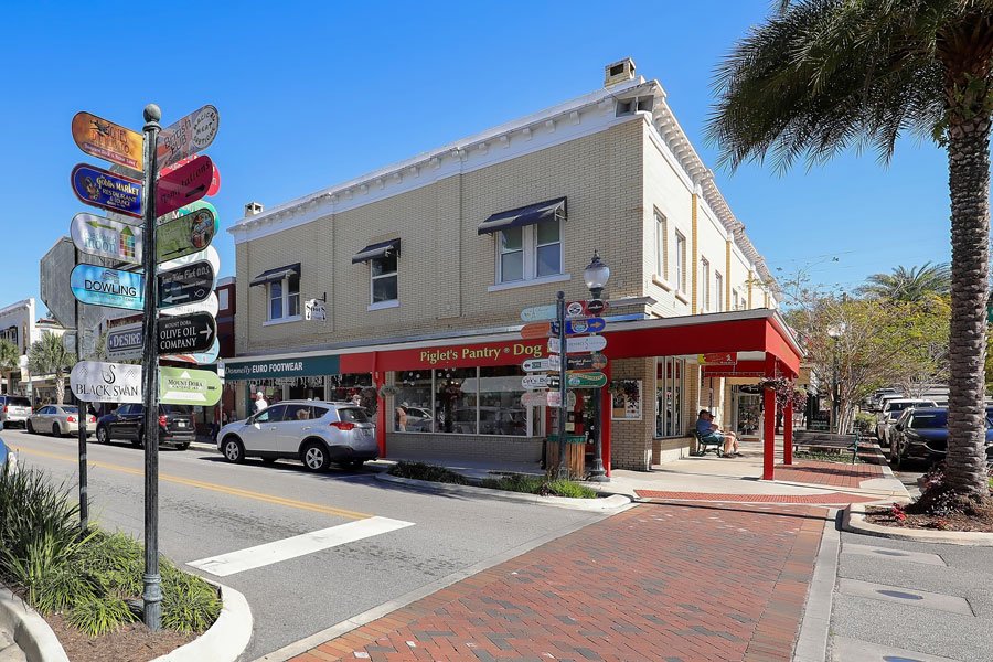 A street-level view of historic downtown Mount Dora, Florida, featuring colorful directional signs, locally owned shops, restaurants and traditional storefronts along a brick-paved pedestrian crossing. Located in Lake County northwest of Orlando, Mount Dora is a popular Central Florida destination known for its antique shops, art festivals, historic architecture and picturesque setting along Lake Dora. Photo credit: Jillian Cain Photography, licensed.