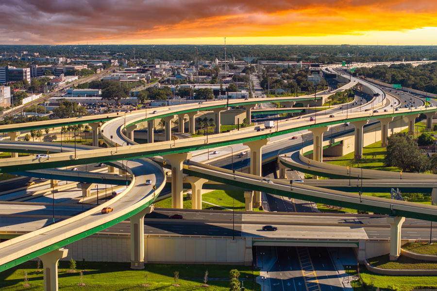 An aerial view of a sprawling multilayer highway interchange in Orlando, Florida, as vehicles travel along elevated ramps and connecting roadways beneath a colorful sunset. The complex network of highways reflects the extensive transportation infrastructure supporting Central Florida's growing communities, tourism industry and regional economy. Photo credit: Bilanol, licensed.