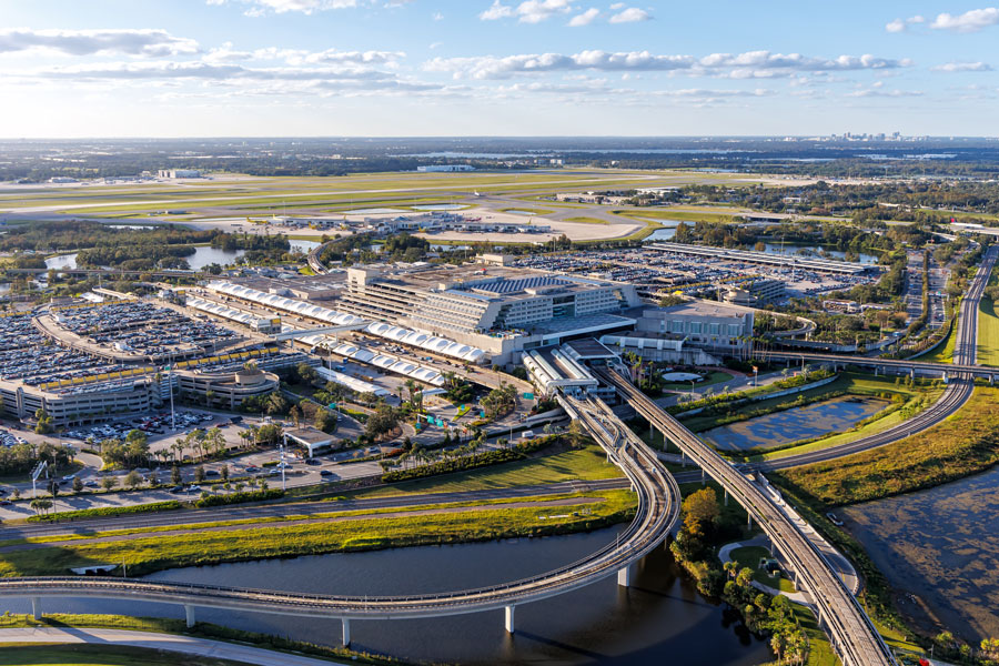 An aerial view of Orlando International Airport in Orlando, Florida, showing its passenger terminal complex, parking facilities, elevated roadways, automated people mover tracks and surrounding runways. As one of the nation's busiest airports, Orlando International Airport serves as a major gateway to Central Florida, connecting millions of travelers each year with the region's theme parks, convention facilities, resorts and surrounding communities. Photo credit: Markus Mainka, licensed.
