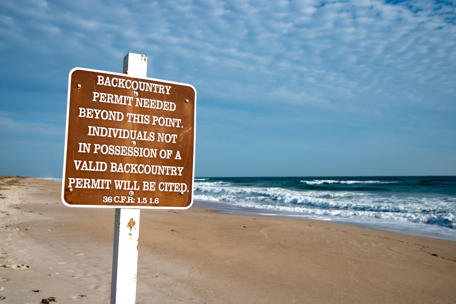 A backcountry access sign marks a protected section of beach at Canaveral National Seashore, where permits are required for travel beyond designated public-use areas. The national seashore preserves a long stretch of undeveloped Atlantic coastline, along with dunes, lagoons, wetlands and wildlife habitat along Florida’s east coast. Visitors exploring the more remote portions of the park should observe posted access restrictions and permit requirements designed to protect sensitive natural areas. Photo credit: Jesse Kunerth; licensed.
